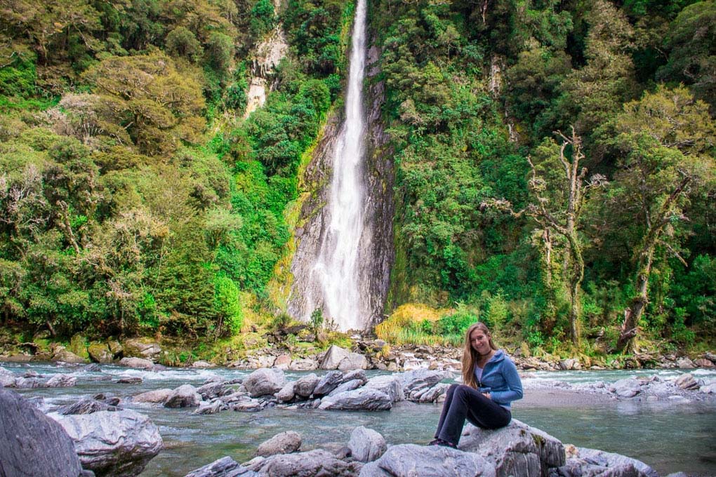 A woman sits on a rock in front of Thunder Creek Falls in New Zealand