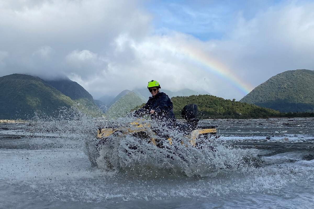 woman driving on a quad bike through a river with mountains behind her