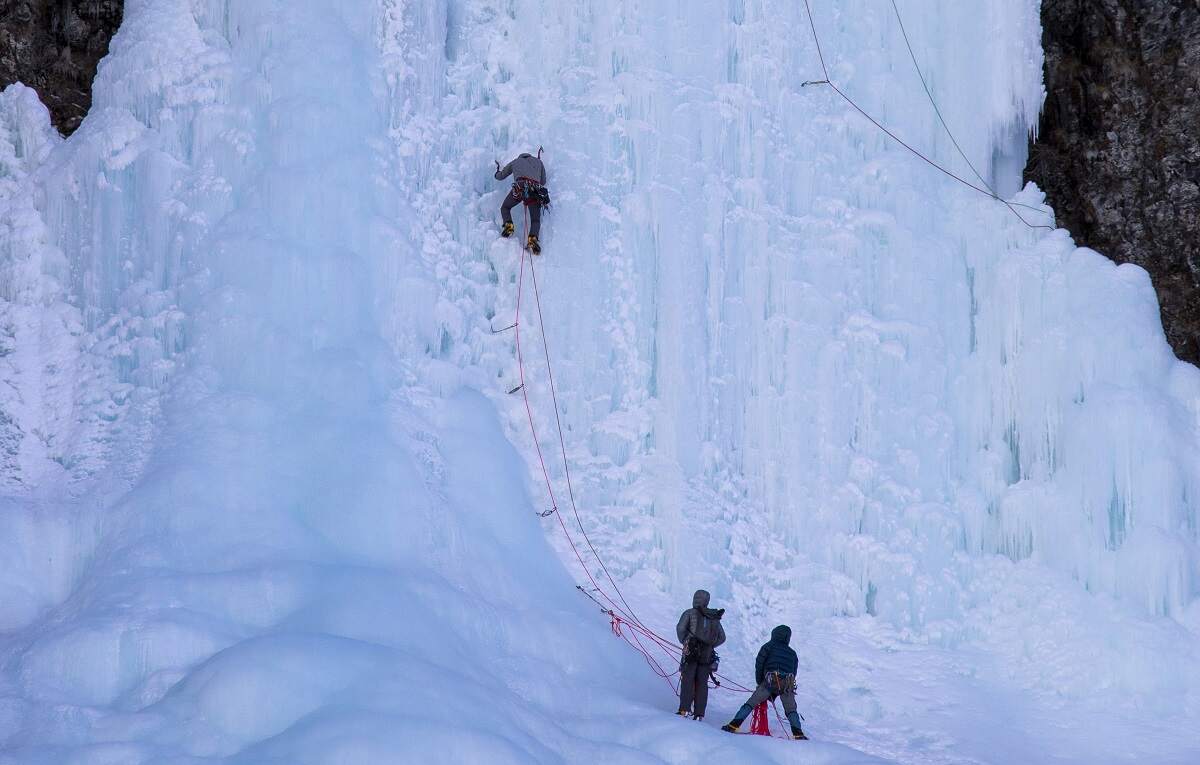 person climbing a frozen waterfall with two people watching from the bottom and feeding a rope in Jasper National Park