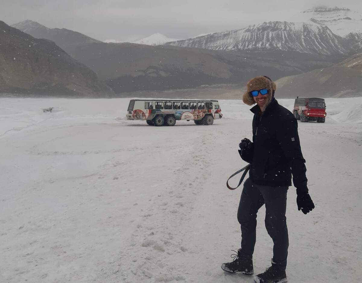 man standign on the Athabasca Glacier in the snow with ICe Explorer vehicles in the background
