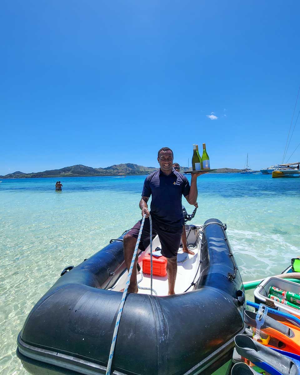 Fijian man holding bottles of wine and smiling ass he stands in a small boat near the sand bar Fiji
