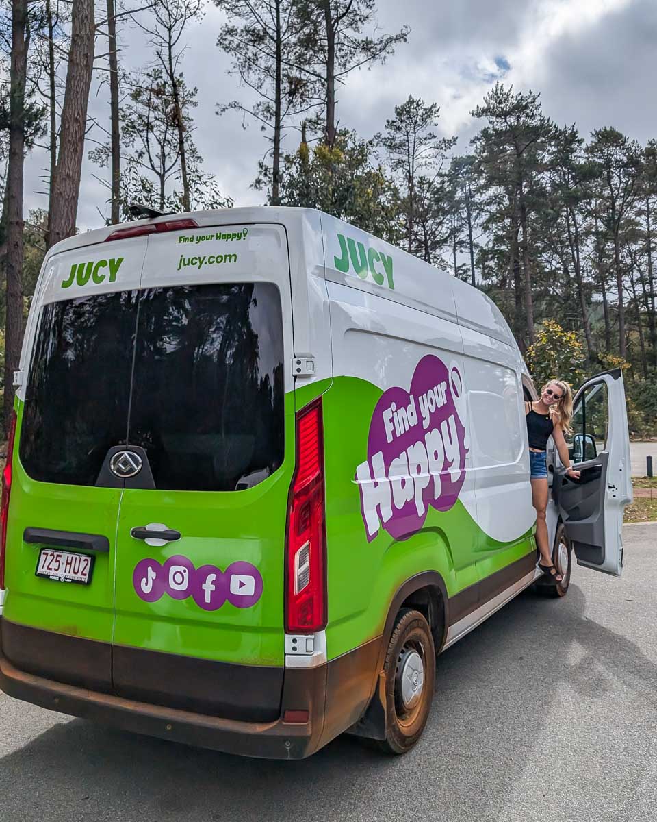 Bailey stands smiles and leans out of a JUCY campervan while camping in Australia